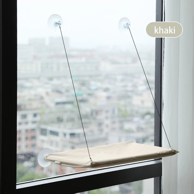 Khaki fabric window hammock suspended by suction cups on high-rise window overlooking city landscape.